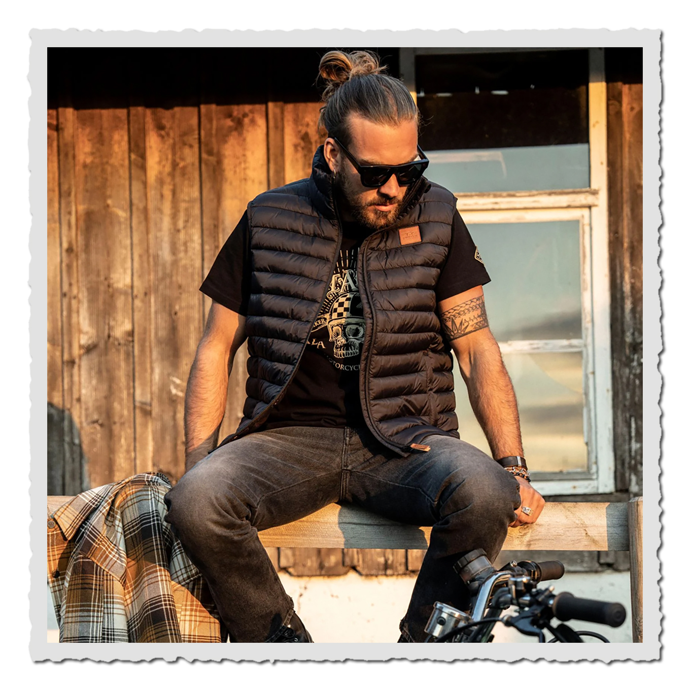 Mann sitzt vor Holzwand, trägt schwarze gesteppte Weste über T-Shirt, Sonnenbrille, Abendlicht.